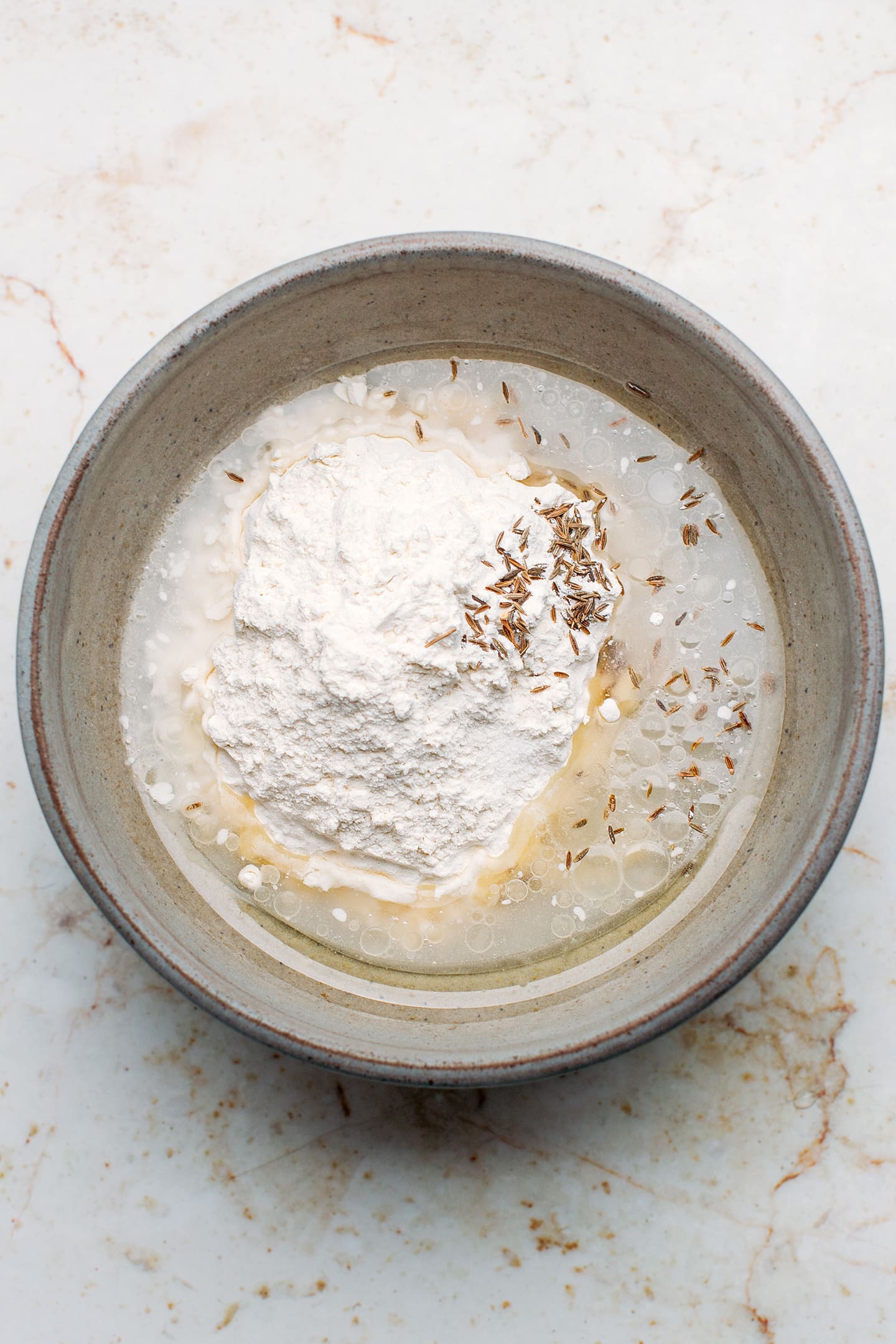 Flour, water, oil, and cumin seeds in a bowl.