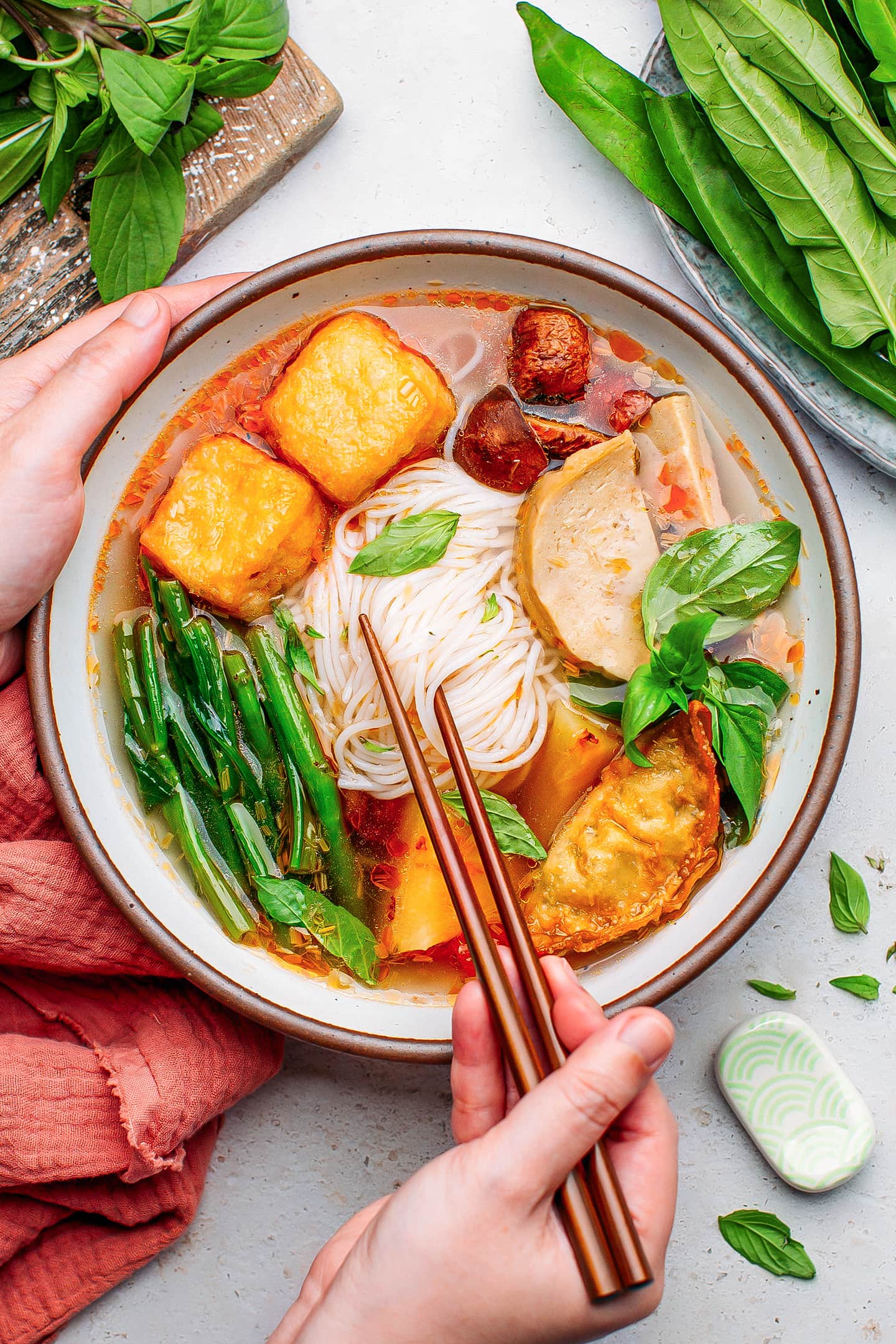 Holding a bowl of hot and sour soup with rice noodles, tofu, and morning glory.