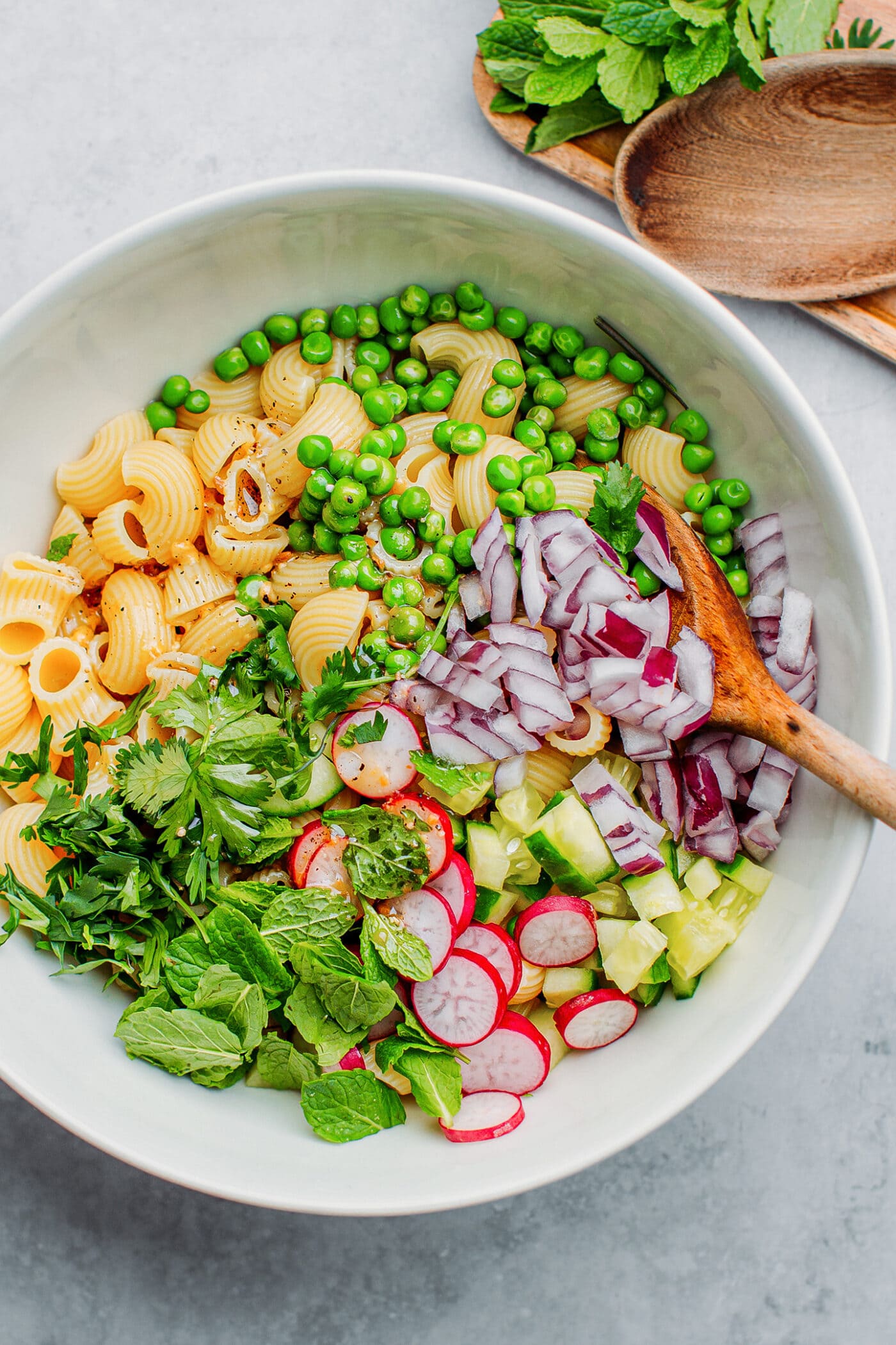 Cooked macaroni, red onions, radishes, cucumber, and fresh herbs in a bowl.